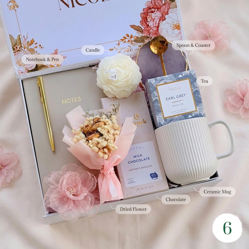 May include: A gift box with a notebook, pen, candle, dried flowers, chocolate, tea, ceramic mug, and a spoon with a coaster. The box is white with floral decorations and a pink ribbon. The mug is white with vertical lines.