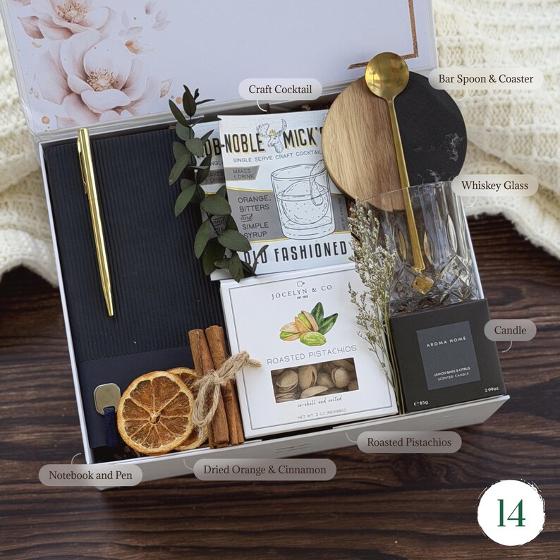 May include: A gift box with a gold pen, notebook, dried orange slices, cinnamon sticks, a craft cocktail kit, roasted pistachios, a whiskey glass, a bar spoon and coaster, and a candle. The box is white with floral accents.