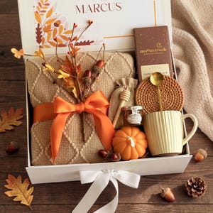 May include: A white gift box containing a tan textured blanket tied with an orange ribbon, a Moonstruck dark chocolate bar, a round woven coaster, a gold spoon, a yellow ribbed mug, a small honey jar with a dipper, and a pumpkin-shaped candle.