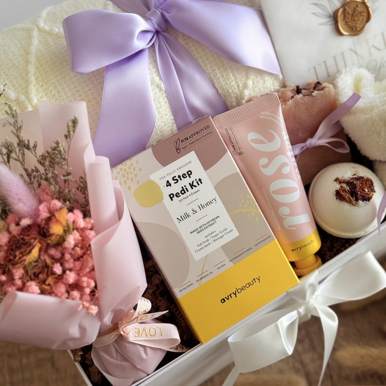 May include: A gift box containing beauty and relaxation products. It features a "4 Step Pedi Kit" box, a pink "rose" cream tube, a bath bomb, soap, and dried flowers. The box is tied with a lilac ribbon and a white ribbon.