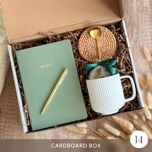 May include: A cardboard gift box containing a sage green notebook with the word NOTES printed in gold, a gold pen, a white ribbed ceramic mug, a small burlap pouch tied with a dark green ribbon, and a round woven rattan coaster with a gold spoon.