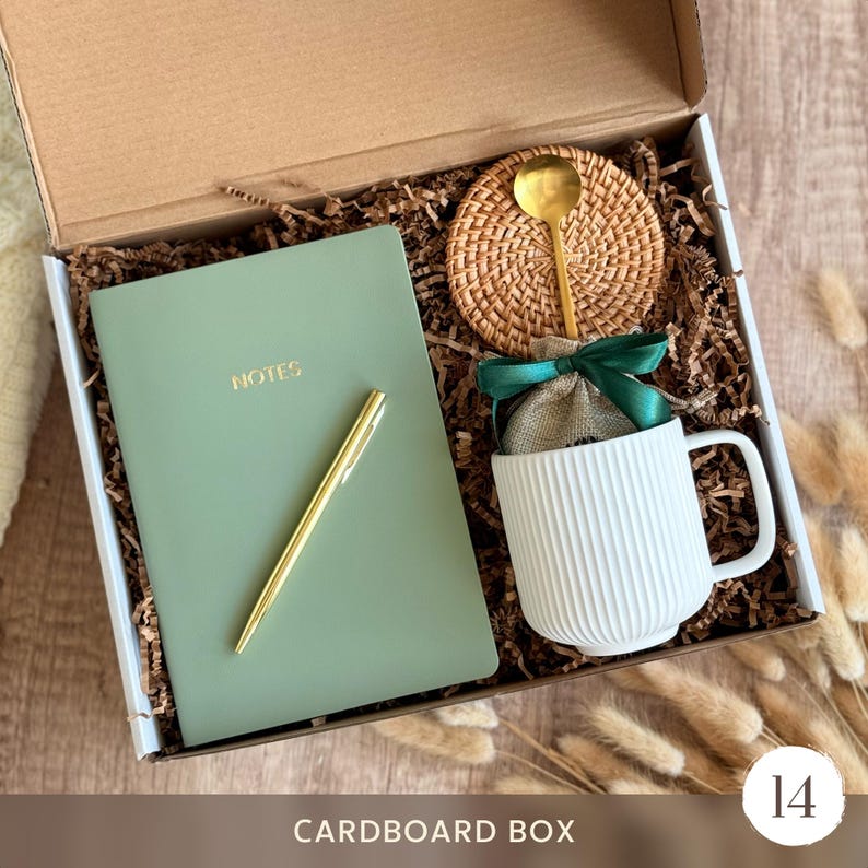 May include: A cardboard gift box containing a sage green notebook with the word NOTES printed in gold, a gold pen, a white ribbed ceramic mug, a small burlap pouch tied with a dark green ribbon, and a round woven rattan coaster with a gold spoon.