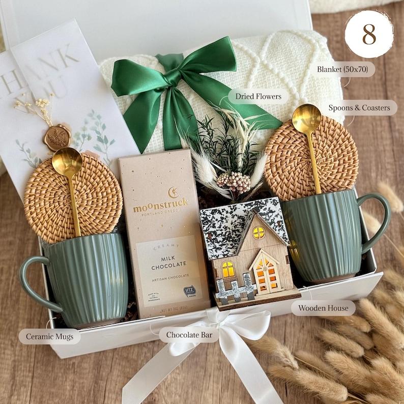 May include: A gift box containing a white knit blanket, two sage green ribbed ceramic mugs with gold spoons and round rattan coasters, a Moonstruck milk chocolate bar, a small wooden house ornament, and a bundle of dried flowers with a thank you card.