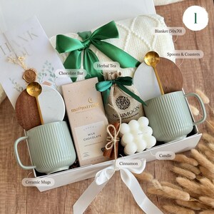 May include: A gift box with a cream-colored blanket, two ribbed ceramic mugs, and a chocolate bar. The box also contains herbal tea, a candle, cinnamon sticks, and gold spoons. The blanket measures approximately 50 x 70 inches.