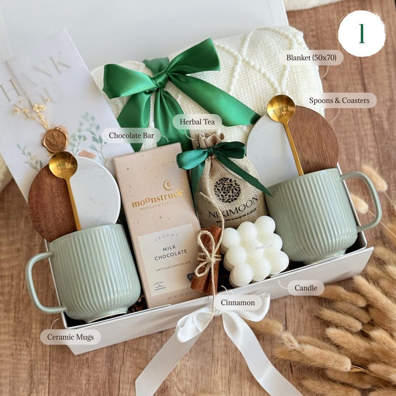 May include: A gift box with a cream-colored blanket, two ribbed ceramic mugs, and a chocolate bar. The box also contains herbal tea, a candle, cinnamon sticks, and gold spoons. The blanket measures approximately 50 x 70 inches.