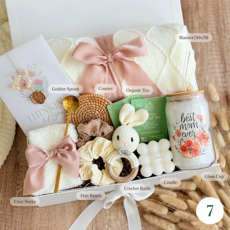 May include: A gift box filled with a variety of items, including a blanket, a glass cup with the text "best mom ever", a crochet rattle, and a card that says "Thinking of You". The box is tied with a white ribbon.