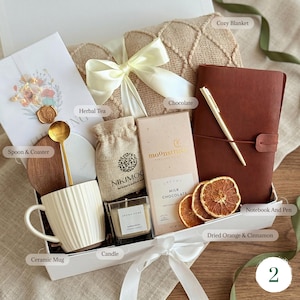 May include: A gift box filled with a cozy blanket, a brown leather notebook and pen, a ceramic mug, a candle, herbal tea, chocolate, a spoon and coaster, and dried orange slices. The box is tied with a white ribbon.