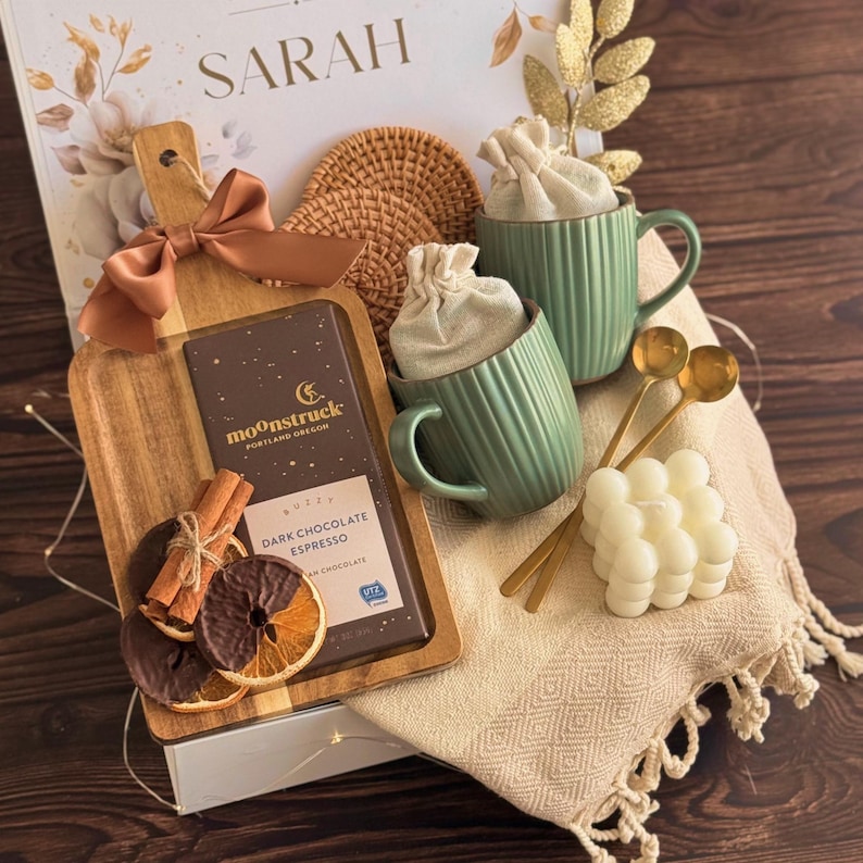 May include: A gift set featuring a wooden serving board with a brown ribbon, a Moonstruck dark chocolate espresso bar, two sage green ribbed mugs with fabric pouches, two gold spoons, a white bubble candle, and a cream-colored tasseled throw blanket.
