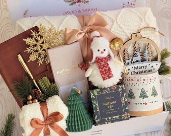 May include: A holiday gift box containing a cream knit blanket with a tan bow, a brown journal, a gold snowflake, a Moonstruck chocolate bar, a plush snowman, a green tree candle, Holiday Tea, and a mug with trees and the text Merry Christmas.