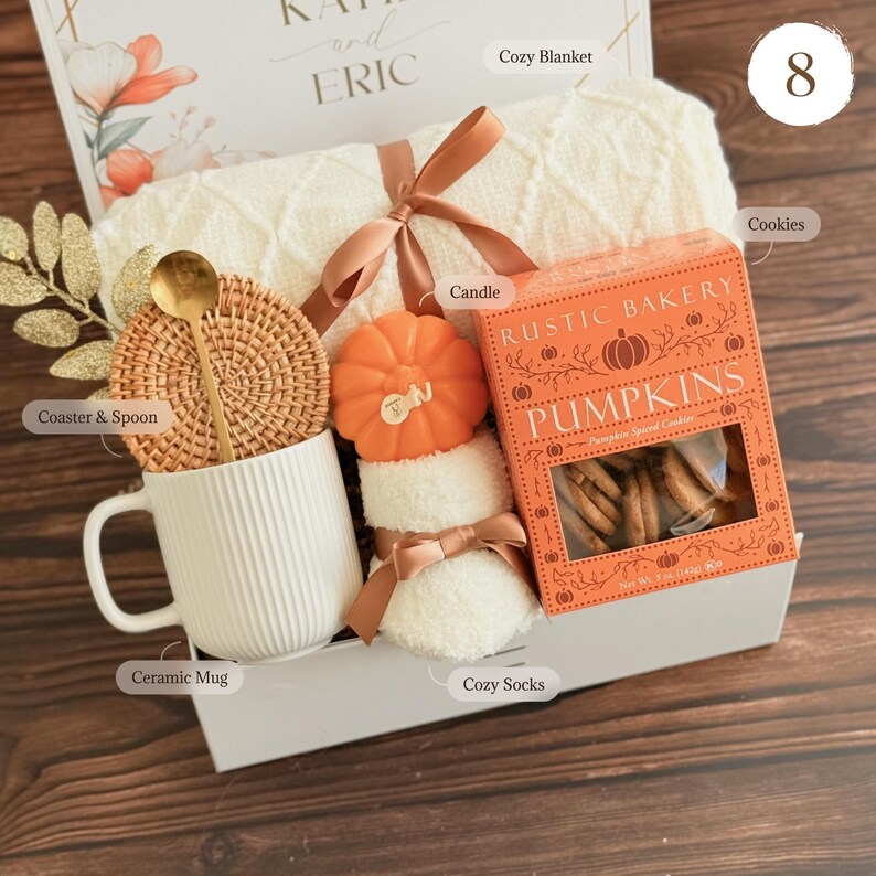 May include: A gift basket with a white ceramic mug, a woven coaster with a gold spoon, a pumpkin-shaped candle, cozy socks, a box of pumpkin spice cookies, and a cream-colored blanket. The box is decorated with the names "KAY" and "ERIC".