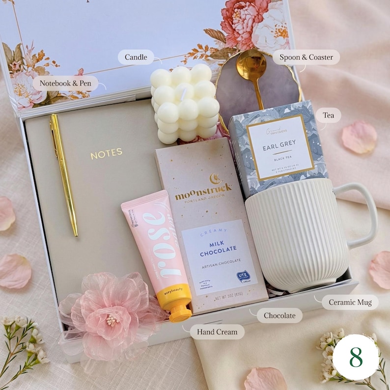 May include: A gift box with a variety of items. Includes a notebook and pen, a candle, a spoon and coaster, tea, a ceramic mug, chocolate, and hand cream. The box is white with floral accents and a pink flower.