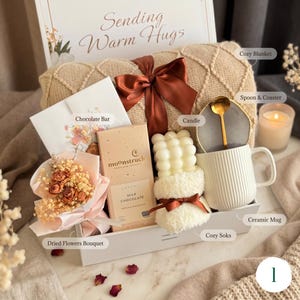 May include: A gift box with a cozy theme, featuring a blanket, chocolate bar, dried flowers, candle, mug, spoon, coaster, and socks. The box is decorated with a ribbon and the text "Sending Warm Hugs".