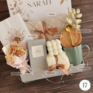 May include: A gift box containing a beige knit blanket tied with a brown ribbon, a box of hot cocoa, a white bubble candle, a pair of fuzzy white socks, a green ceramic mug, a gold spoon, a woven coaster, and a bouquet of dried flowers.