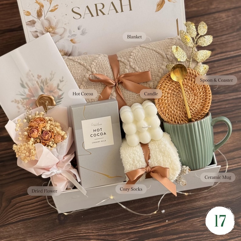 May include: A gift box containing a beige knit blanket tied with a brown ribbon, a box of hot cocoa, a white bubble candle, a pair of fuzzy white socks, a green ceramic mug, a gold spoon, a woven coaster, and a bouquet of dried flowers.
