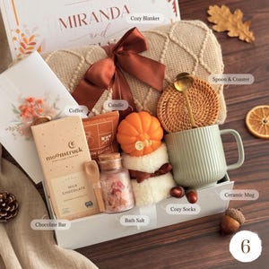 May include: A gift box containing a beige knit blanket with a brown ribbon, a Moonstruck milk chocolate bar, a small jar of bath salts, a pumpkin-shaped candle, a pair of white fuzzy socks, a green ribbed ceramic mug, and a gold spoon with a coaster.