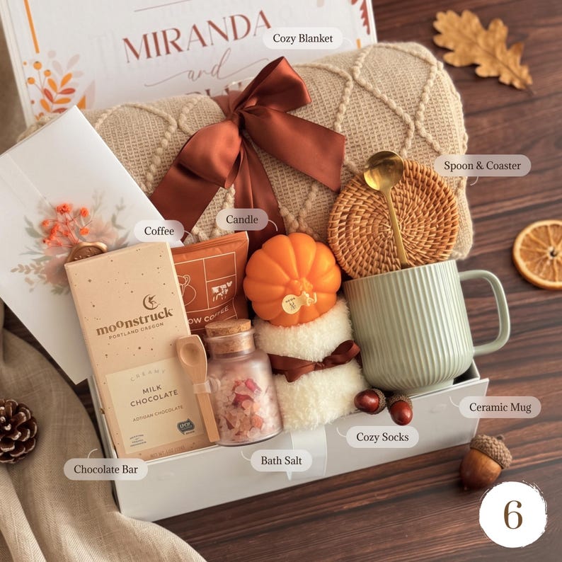May include: A gift box containing a beige knit blanket with a brown ribbon, a Moonstruck milk chocolate bar, a small jar of bath salts, a pumpkin-shaped candle, a pair of white fuzzy socks, a green ribbed ceramic mug, and a gold spoon with a coaster.