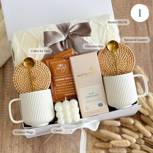 May include: A white gift box filled with cozy items for a relaxing evening. The box includes a white blanket, two ceramic mugs, a candle, a chocolate bar, coffee for two, and two spoons with coasters.