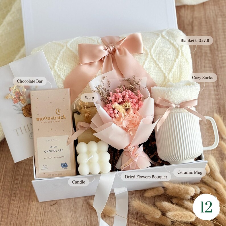 May include: A gift box filled with a cream-colored blanket (50x70 inches), cozy socks, a ceramic mug, a chocolate bar, soap, a candle, and a dried flower bouquet. The box is tied with a pink ribbon.