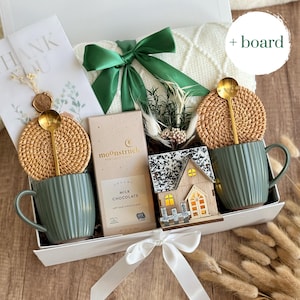 May include: A gift box with a green ribbon, containing two green mugs with gold spoons, woven coasters, a chocolate bar, a decorative wooden house, and a white blanket. A card with the words "THANK YOU" is also included.