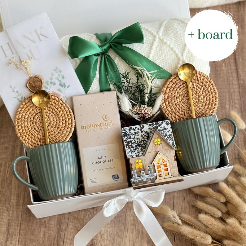 May include: A gift box with a green ribbon, containing two green mugs with gold spoons, woven coasters, a chocolate bar, a decorative wooden house, and a white blanket. A card with the words "THANK YOU" is also included.