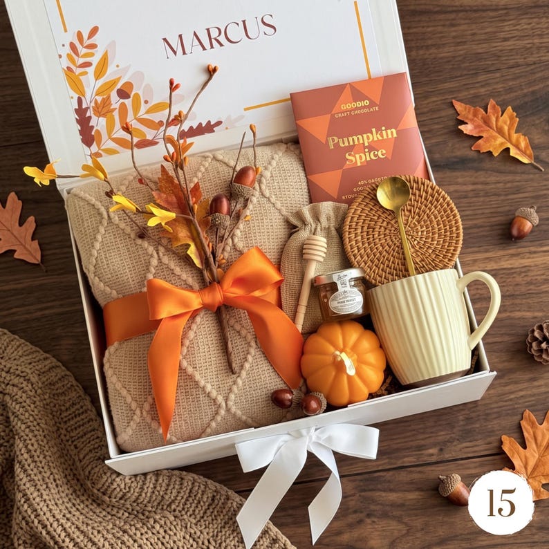 May include: A gift box containing a beige knit blanket tied with an orange ribbon, a Goodio Pumpkin Spice chocolate bar, a small jar of honey with a wooden dipper, a pumpkin-shaped candle, a round woven coaster, and a ribbed cream mug with a gold spoon.