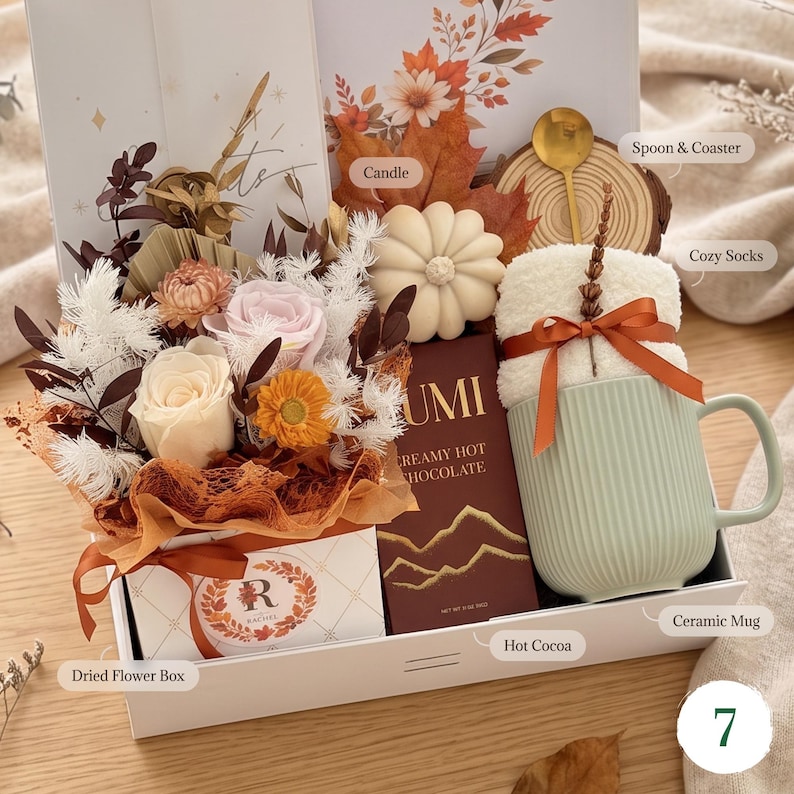 May include: A gift box containing a dried flower arrangement in a box labeled R, a pumpkin-shaped candle, a wooden coaster with a gold spoon, a pair of white fuzzy socks, a box of Lumi creamy hot chocolate, and a ribbed light green ceramic mug.