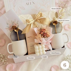 May include: A gift box with a white blanket, two white ceramic mugs, chocolate, a candle, dried flowers, and gold spoons. The box is decorated with a gold bow and floral accents. The text "ALEX" is visible.