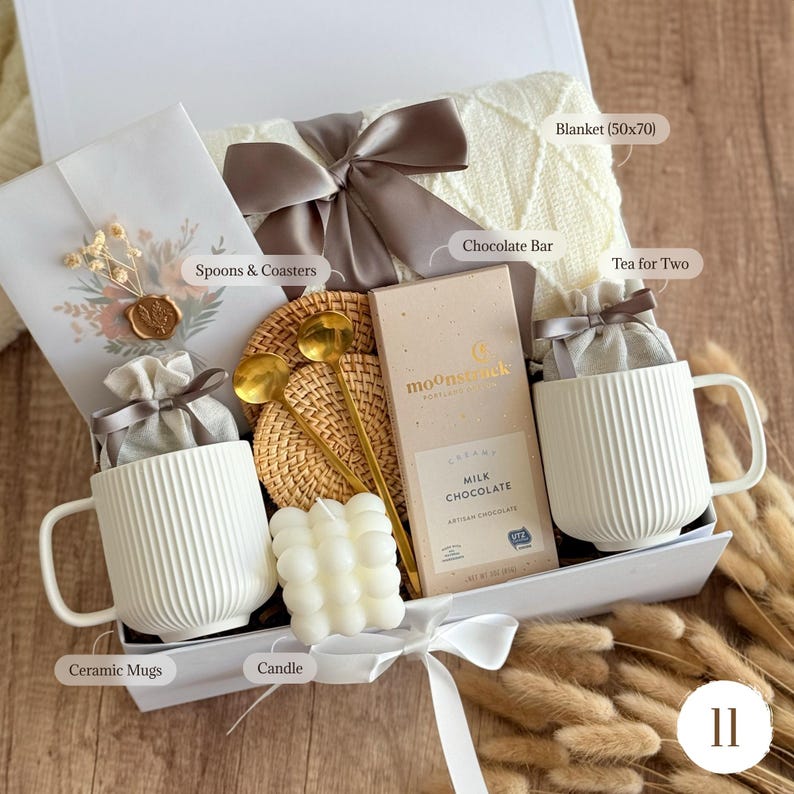 May include: A gift box with a white blanket (50x70 inches), ceramic mugs, a chocolate bar, tea, a candle, spoons, and coasters. The box is tied with a ribbon. The items are arranged on a white tray.