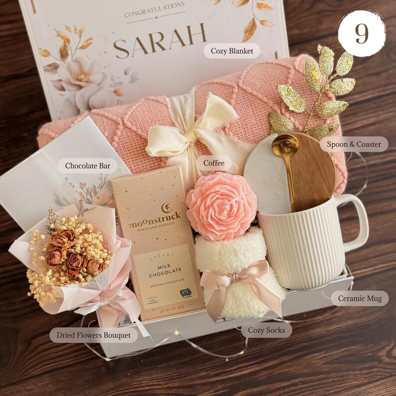 May include: A gift box with a cozy blanket, ceramic mug, chocolate bar, dried flowers, and other items. The box includes a pink blanket, a white mug, and a chocolate bar with the text "moonstruck". The box is decorated with a bow and a gold spoon.