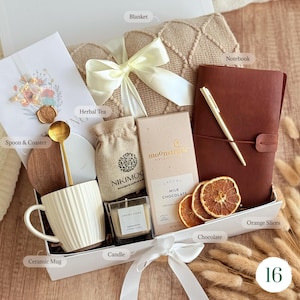 May include: A gift box containing a beige textured blanket with a white ribbon, a brown notebook with a gold pen, a white ribbed ceramic mug, a gold spoon, a wooden coaster, a small burlap bag, a candle, milk chocolate, and dried orange slices.