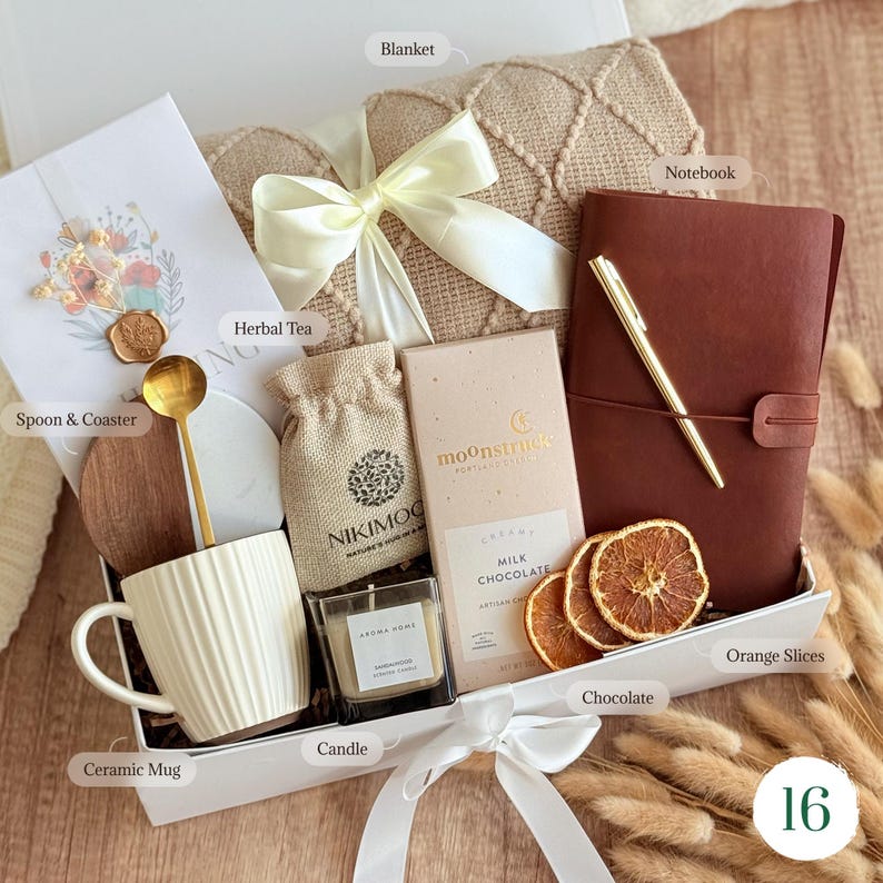 May include: A gift box containing a beige textured blanket with a white ribbon, a brown notebook with a gold pen, a white ribbed ceramic mug, a gold spoon, a wooden coaster, a small burlap bag, a candle, milk chocolate, and dried orange slices.