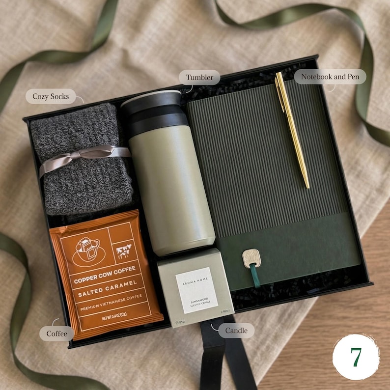 May include: A gift box with a variety of items, including a gray tumbler, cozy socks, a notebook with a gold pen, a candle, and a packet of coffee. The box is black with a green ribbon and is arranged on a neutral-colored surface.
