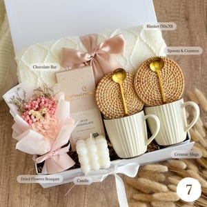 May include: A gift box containing a cream-colored blanket (50x70 inches), two ceramic mugs, a chocolate bar, a dried flower bouquet, a candle, two woven coasters with gold spoons, and a pink bow.