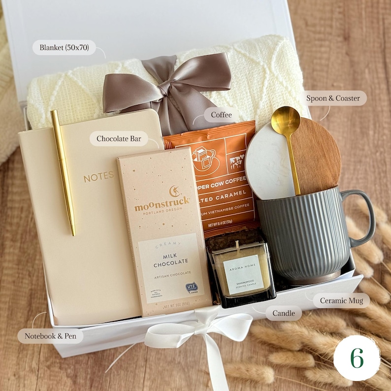 May include: A gift box filled with a cream-colored blanket, a notebook and pen, a chocolate bar, coffee, a ceramic mug with a spoon and coaster, and a candle. The blanket measures 50x70 inches. The box is tied with a ribbon.