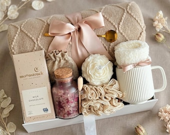 May include: A gift set containing a beige textured throw blanket tied with a satin ribbon, a Moonstruck milk chocolate bar, a glass jar of pink bath salts with a cork lid, a white flower-shaped candle, a beige loofah, and a white ribbed mug.