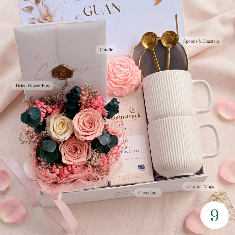 May include: A gift box with a dried flower arrangement, two white ceramic mugs, a chocolate bar, a pink rose-shaped candle, and gold spoons. The box contains a bouquet of pink and cream roses.