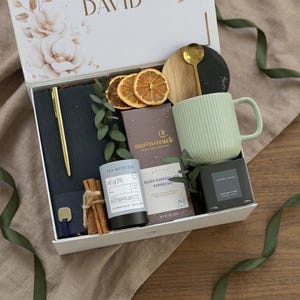 May include: A gift box with a variety of items, including a gold pen, a notebook, a mug, tea, chocolate, and a candle. The mug is light green and has vertical lines. The box is white with floral accents and the name "DAVID" on the lid.