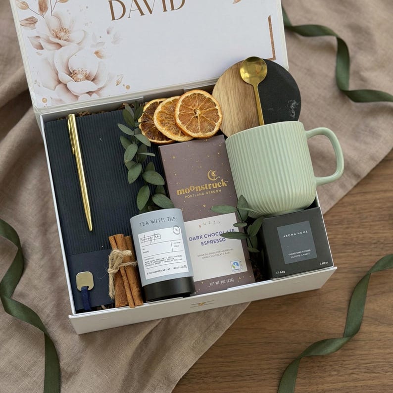 May include: A gift box with a variety of items, including a gold pen, a notebook, a mug, tea, chocolate, and a candle. The mug is light green and has vertical lines. The box is white with floral accents and the name "DAVID" on the lid.