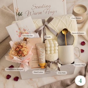 May include: A gift basket with a cozy theme, featuring a cream-colored blanket, a ceramic mug, a candle, and a bouquet of dried flowers. The basket also includes a chocolate bar, hand lotion, and cozy socks, all tied together with a ribbon.