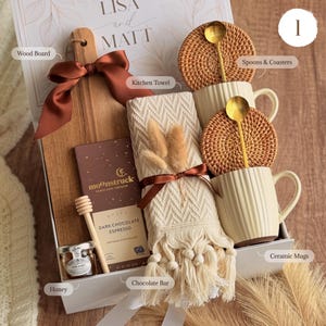 May include: A gift box containing a wooden serving board with a rust-colored ribbon, a Moonstruck dark chocolate espresso bar, a small jar of honey with a wooden dipper, a cream-colored kitchen towel, two ribbed ceramic mugs, and two rattan coasters.