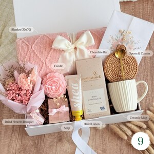 May include: A gift box filled with a pink blanket (50x70 inches), a dried flower bouquet, a rose-shaped candle, a bar of soap, hand lotion, a chocolate bar, a ceramic mug, a spoon, and a coaster. The box is tied with a white ribbon.