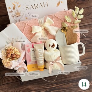 May include: A gift box with a pink blanket, bath bomb, ceramic mug, cozy socks, dried flowers, skin care kit, and hand cream. The box is decorated with a bow and gold accents. The box is labeled "SARAH".