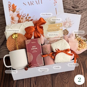 May include: A gift box containing a white ribbed ceramic mug, a gold spoon, a woven coaster, a box of hot cocoa, a pink quartz roller, a shea butter hand mask, a ribbed pink candle, white fuzzy socks, and a small bouquet of dried flowers.