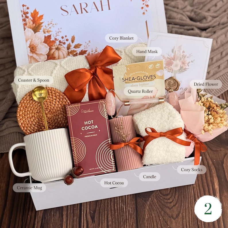 May include: A gift box containing a white ribbed ceramic mug, a gold spoon, a woven coaster, a box of hot cocoa, a pink quartz roller, a shea butter hand mask, a ribbed pink candle, white fuzzy socks, and a small bouquet of dried flowers.