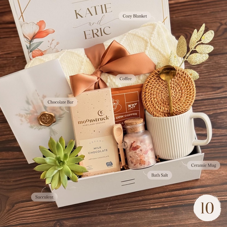 May include: A gift box with a cozy cream-colored blanket, coffee, chocolate, bath salts, a ceramic mug, and a succulent. The box is tied with a brown ribbon and includes a card that reads "KATIE and ERIC".
