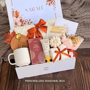 May include: A white gift box containing a white ribbed mug with a gold spoon, a box of hot cocoa, a tube of shea butter, a pair of shea gloves, a white pumpkin-shaped candle, a small dried flower bouquet, and a white knit item with a rust-colored bow.