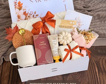 May include: A white gift box containing a white ribbed mug with a gold spoon, a box of hot cocoa, a tube of shea butter, a pair of shea gloves, a white pumpkin-shaped candle, a small dried flower bouquet, and a white knit item with a rust-colored bow.