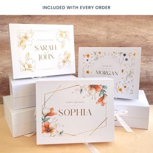 May include: White gift boxes with floral designs and text. One box reads "Sarah and John" with floral accents. Another says "Morgan" with a floral border. A third box says "Sophia" with orange flowers and a gold geometric frame.