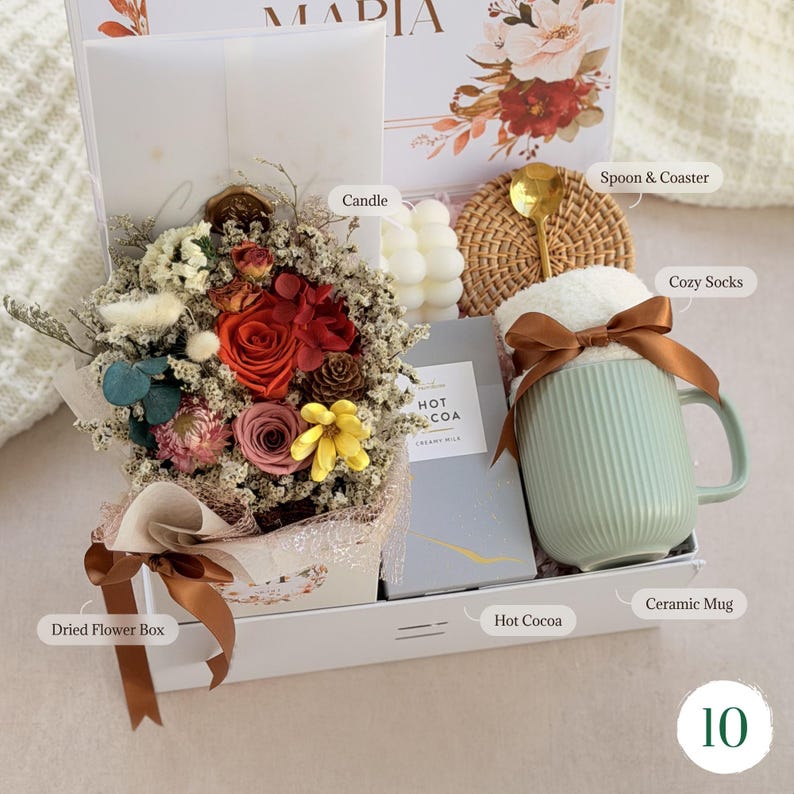 May include: A gift box with a dried flower arrangement, a light blue ceramic mug, cozy socks, hot cocoa, a candle, and a spoon on a coaster. The dried flower arrangement includes red, yellow, and pink flowers. The box is white with a brown ribbon.