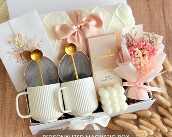 May include: A gift box with a white lid, containing two white ribbed mugs with gold spoons, coasters, a small bouquet of pink flowers, chocolate, and a cream-colored blanket tied with a pink bow. The box is labeled "PERSONALIZED MAGNETIC BOX".